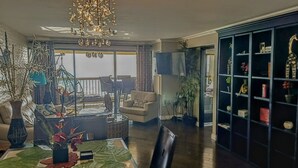 A spacious living room with a large chandelier, a dining area with a table set for two, and a black shelving unit filled with decorative items. - Direct Oceanfront in the Heart of Waikiki! Spectacular New Custom Reno! (Honolulu)