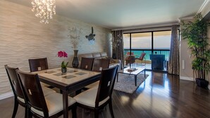 A dining area with a table and chairs, a chandelier, a vase with flowers, and a view of the ocean through large windows. - Direct Oceanfront in the Heart of Waikiki! Spectacular New Custom Reno! (Honolulu)