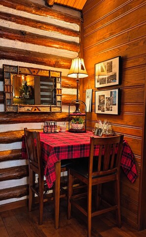 Interior de una cabaña de madera con una mesa de comedor dispuesta para cuatro personas, una lámpara y cuadros en la pared.