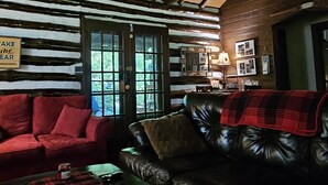 Interior de una cabaña de troncos con un sofá de cuero rojo y negro, una mesa de madera y una chimenea.