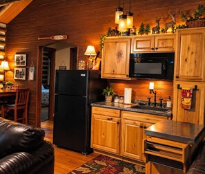 A cozy kitchen with wooden cabinets, a black refrigerator, and a microwave. There is a dining area with a table and chairs, and a living room with a leather couch.