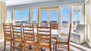 Un comedor con sillas de madera y una mesa, ventanas amplias con vistas, y un sillón gris.