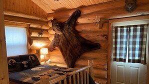 Une chambre rustique en rondins de bois, avec une grande tête d’ours fixée au mur, un lit en bois recouvert d’une couverture à carreaux, une lampe et une porte ornée de rideaux à carreaux.