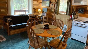 L’intérieur chaleureux d’un chalet avec une table à manger en bois entourée de chaises, un canapé, un téléviseur, une bibliothèque et un coin cuisine équipé d’une cuisinière et d’un four à micro-ondes.
