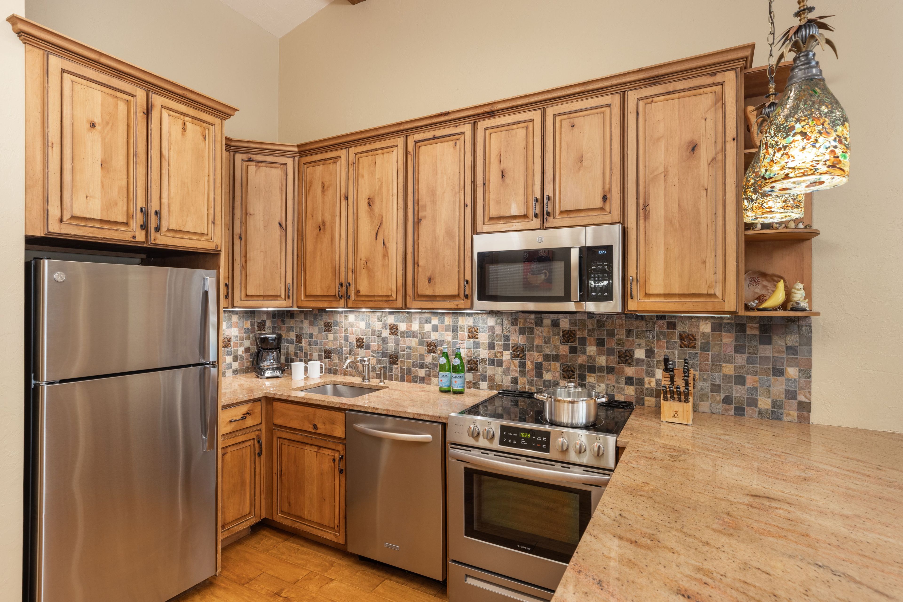 A modern kitchen with wooden cabinets, stainless steel appliances, a tiled backsplash, and a hanging light fixture.