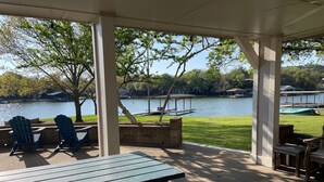 A covered outdoor seating area with Adirondack chairs and a picnic table, overlooking a lake with boats and a dock. - Granbury Lake Rustic Retreat on Quiet Cove (Granbury)