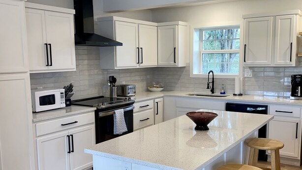 A modern kitchen with white cabinetry, a central island with stools, a stainless steel trash can, and a window providing natural light. - Granbury Lake Rustic Retreat on Quiet Cove (Granbury)
