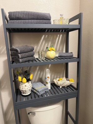 A multi-tiered metal shelf with towels, a vase with yellow flowers, and a bottle of lotion. - Tastefully upgraded. One of the only resort units with a full size laundry team. (Kihei)