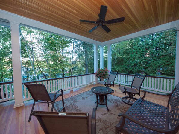 A covered porch with wooden flooring, white railings, and a ceiling fan. There are several chairs and a table with a floral table runner.