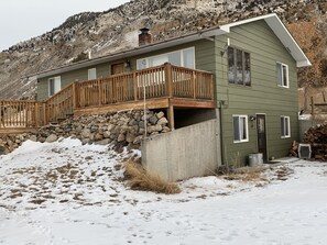 A two-story house with a wooden deck, stone foundation, and a mountainous backdrop.