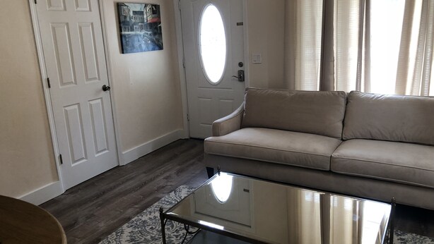 Een woonkamer met een beige bank, een glazen salontafel, een deur met een raam en een kamerbreed tapijt met een patroon.