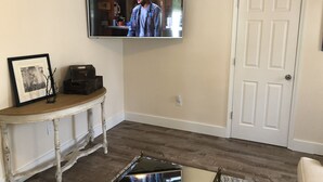 Een woonkamer met een aan de muur bevestigde televisie, een houten tafel met een fotolijstje en een klein beeldje, en een witte deur.