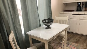Een kleine, witte eettafel met een zwarte decoratieve schaal in het midden, omringd door witte stoelen. Achtergrond toont een keuken met witte kastjes en een koffiezetapparaat.