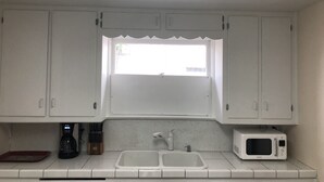 Een keuken met witte kastjes, een raam boven de spoelbak, een koffiezetapparaat en een magnetron.