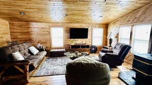 Ein gemütliches Blockhaus-Zimmer mit Holzwänden und Holzböden, einem Wand-Fernseher, einer Couch, einem Sessel, einem kleinen Couchtisch und einem Kamin.