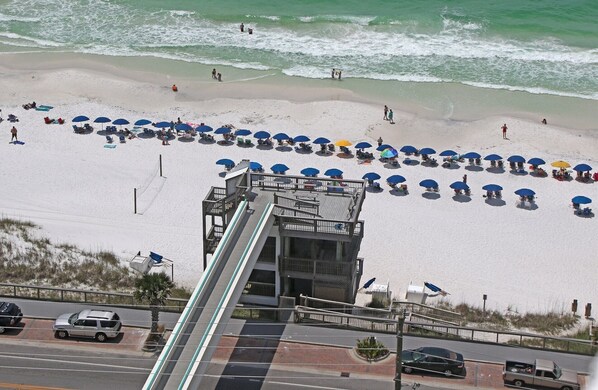Une plage bordée de rangées de parasols et de chaises, une tour de surveillance pour les sauveteurs, et une route où circulent des voitures.