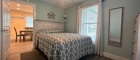 A bedroom with a bed, a ceiling fan, a window with blinds, a bedside lamp, and a door leading to a dining area.