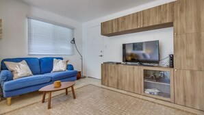 Un salon avec un canapé bleu, une table basse, un téléviseur posé sur un meuble en bois et une fenêtre avec des stores.