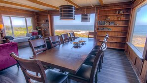Uma sala de jantar com uma mesa e cadeiras de madeira, uma estante e vista para o oceano.
