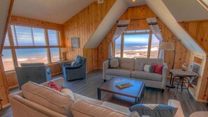 Quarto acolhedor com uma grande janela que oferece vista para a praia, um sofá, uma cadeira, uma mesa pequena e um abajur.