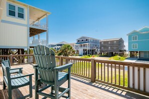 A wooden deck with Adirondack chairs, a wooden railing, and a view of multi-story buildings. - Coastal Breeze: Home Close to Beach Access and Town Park (Oak Island)