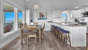 A modern kitchen with white cabinetry, a central island with bar stools, and a dining area with wooden chairs. - Spinnaker: Oceanfront Home with Expansive Views (Oak Island)