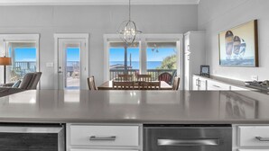 A modern kitchen with a large island, white cabinetry, and a view of the ocean through large windows. - Spinnaker: Oceanfront Home with Expansive Views (Oak Island)
