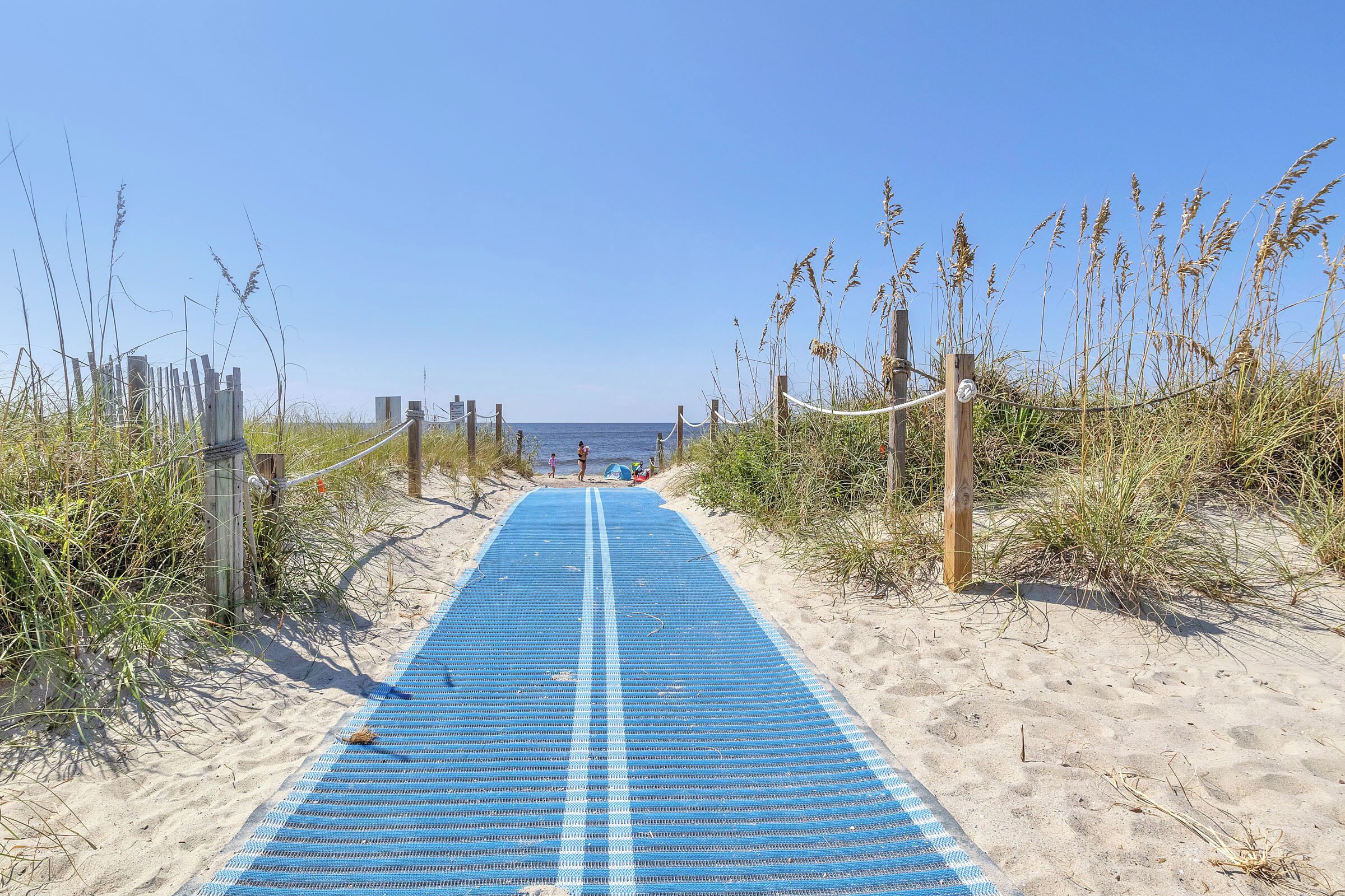 A sandy beach path with a blue mat leading to the sea, surrounded by tall grasses and wooden posts.