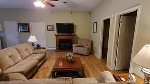 Una sala de estar con un sofá color beige, una mesa de centro de madera, una chimenea, un televisor y una puerta que da a otra habitación.