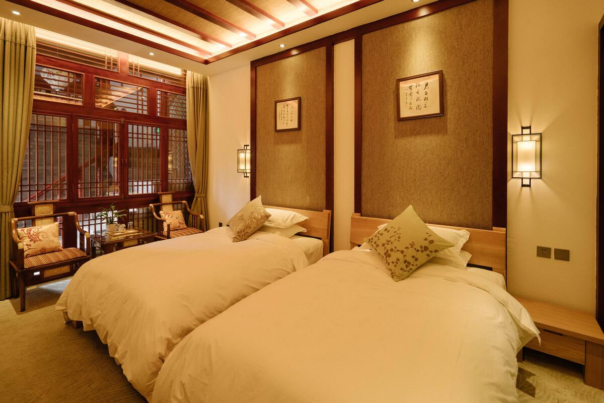 Quarto de hotel com duas camas, cabeceiras de madeira, uma janela grande com cortinas, uma mesinha com uma planta e duas luminárias fixadas na parede.