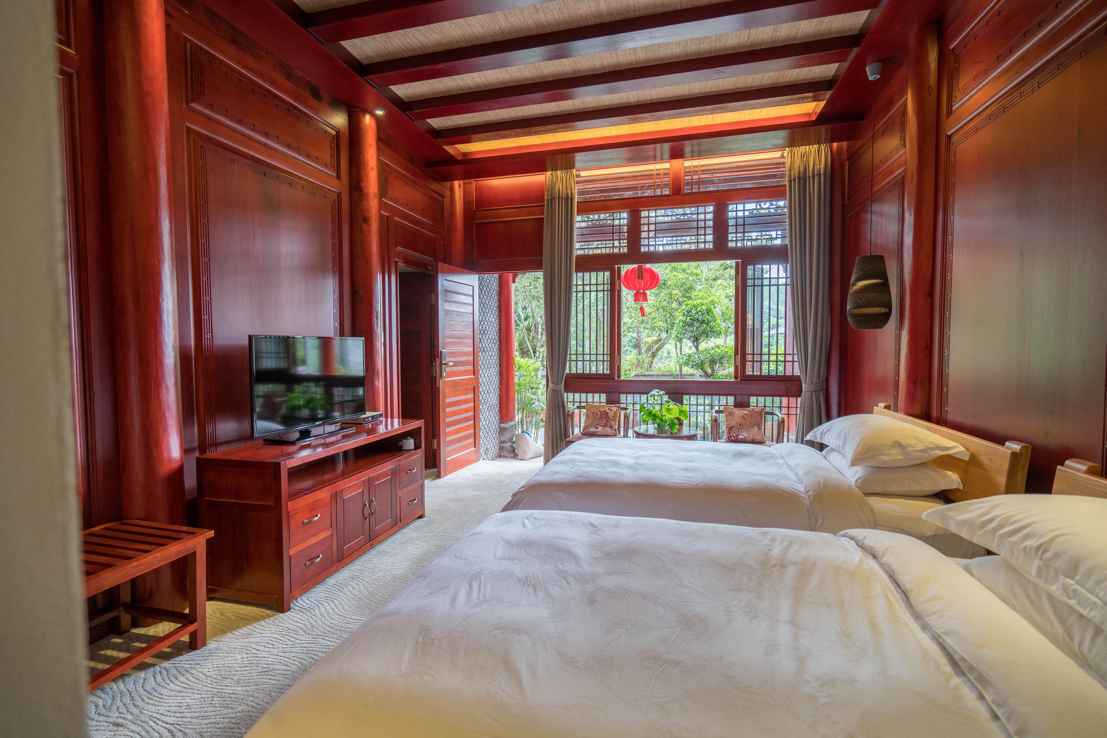 A traditional room with two beds, a television on a wooden cabinet, and a view of greenery outside.