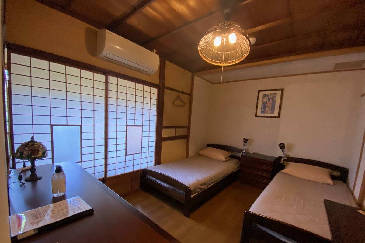 Una habitación tradicional japonesa con suelo de tatami, techo de madera bajo y puertas correderas shoji. Hay dos camas con estructura de madera y ropa de cama blanca, una mesita con una lámpara y un aire acondicionado fijado a la pared.