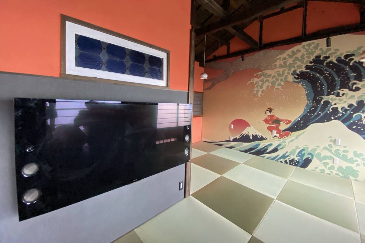 Una habitación con una televisión de pantalla grande y plana montada en una pared gris, una obra de arte enmarcada encima y una pintura tradicional japonesa de olas en la pared contigua.