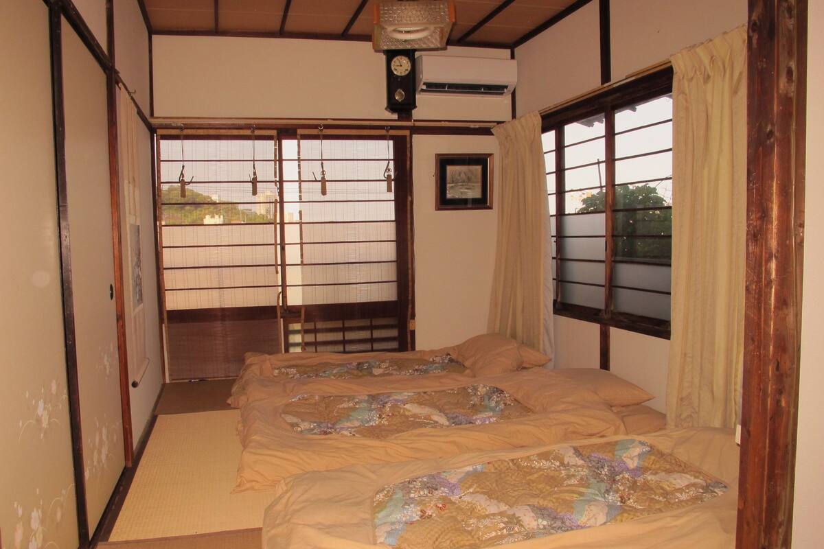 Una habitación tradicional japonesa con suelo de tatami, una cama con ropa de cama floral, una puerta corredera con vistas y un equipo de aire acondicionado.