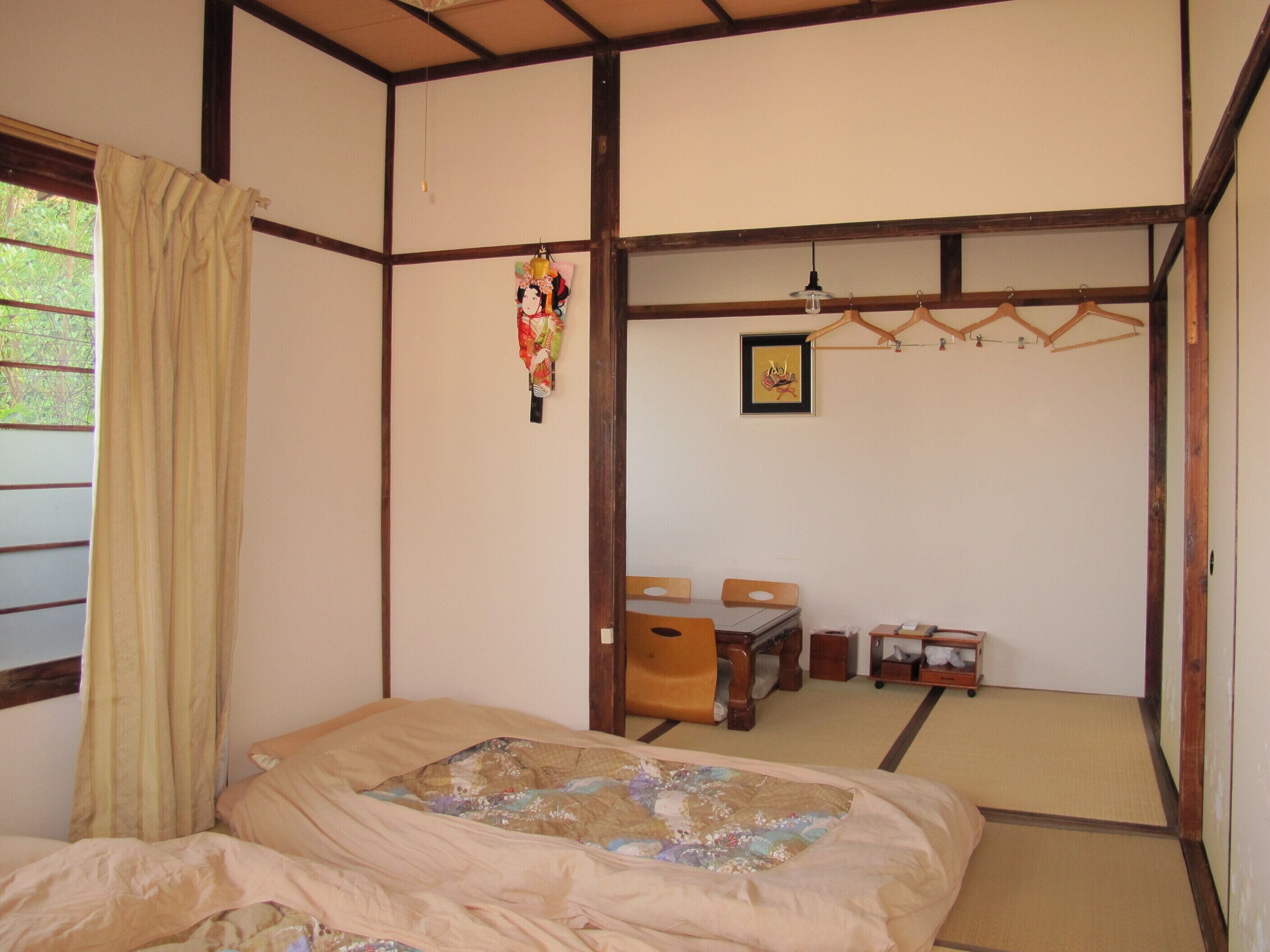 Una habitación japonesa tradicional con suelo de tatami, una mesa de madera baja con sillas, una cama con colcha y una pantalla shoji colgante.
