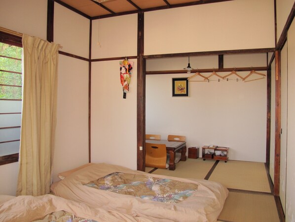 A traditional Japanese room with tatami flooring, a low wooden table with chairs, a bed with a quilt, and a hanging shoji screen.