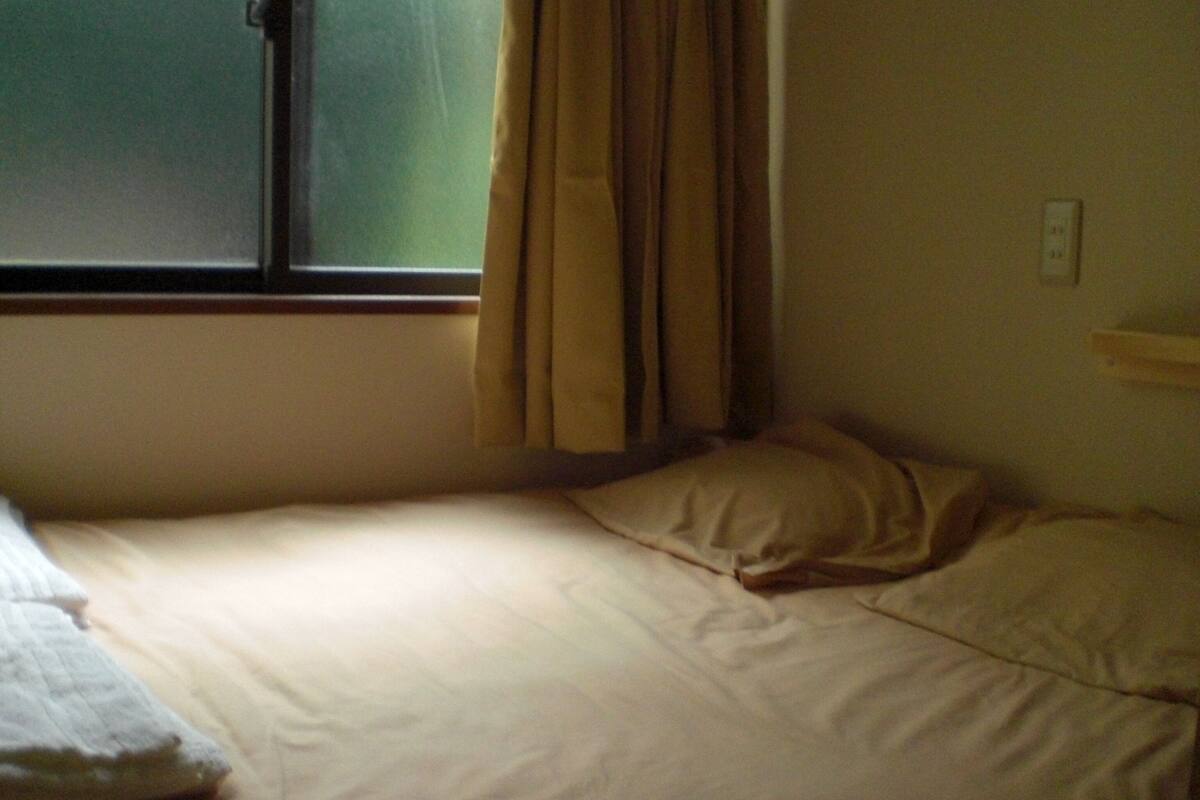 Una cama individual con una colcha blanca y dos almohadas, una ventana con cortinas y un enchufe de pared.