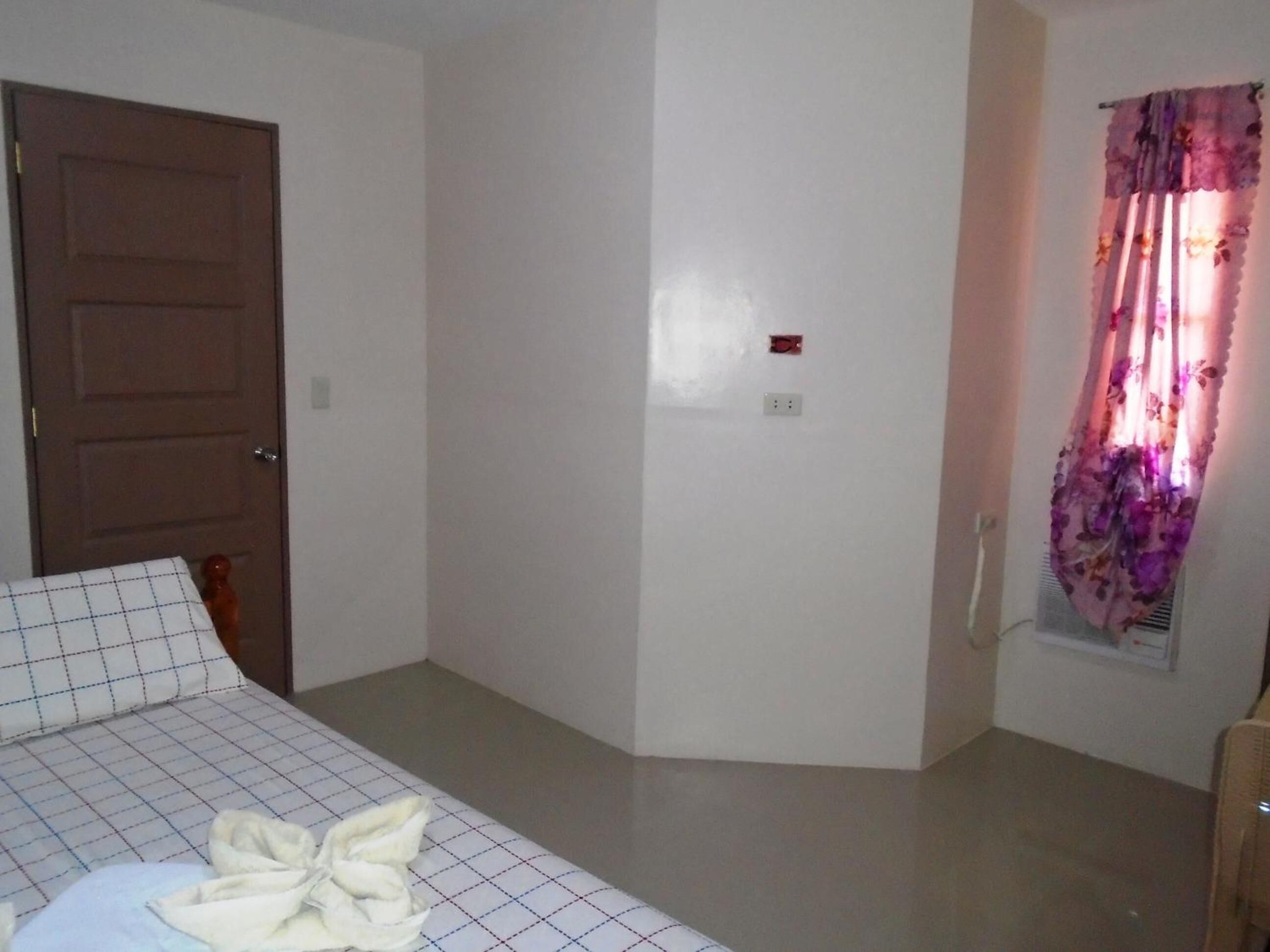 A bedroom with a bed, a door, a power outlet, and a window with floral curtains.