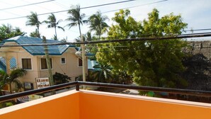 Ein Balkon mit einer orangefarbenen Wand und einem Metallgeländer. Im Hintergrund sind Gebäude mit blauen Dächern und Palmen zu sehen.