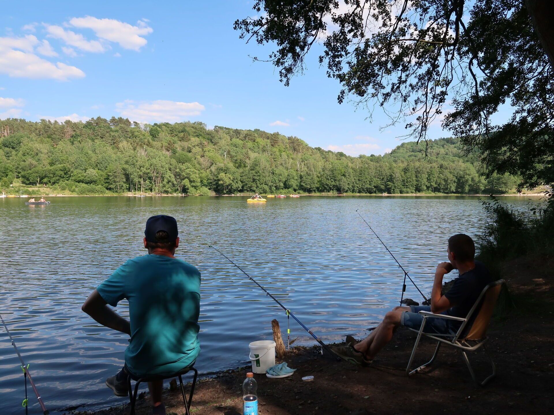 Dues persones pescant a un llac, amb vaixells al fons.