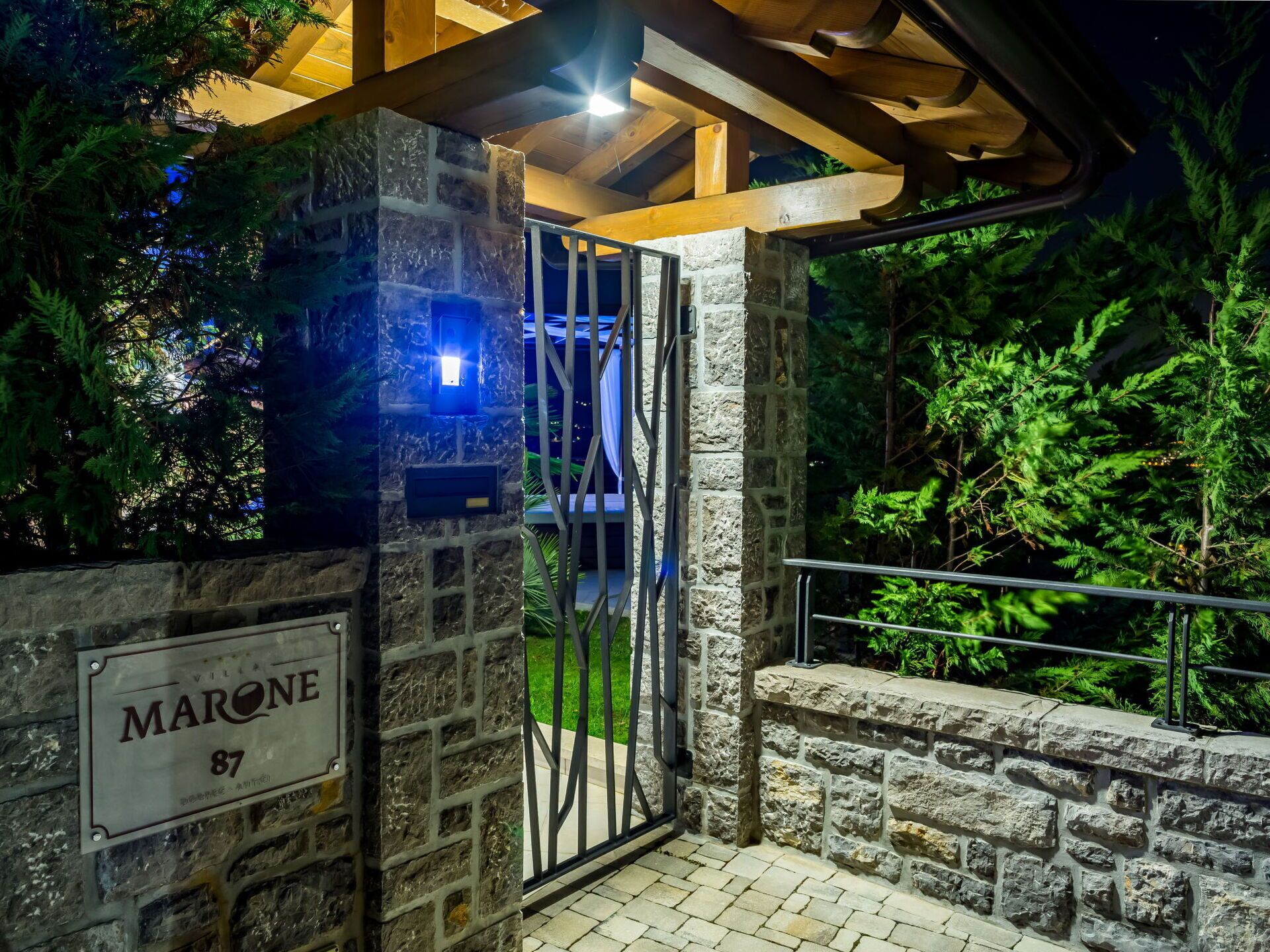 A stone entrance with a metal gate, a sign reading "MARONE 87", and a wooden structure above.
