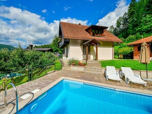 Un espace aménagé au bord d’une piscine à l’eau d’un bleu limpide, avec des marches en pierre menant à une maison dont le toit est recouvert de tuiles rouges, et des chaises longues blanches.