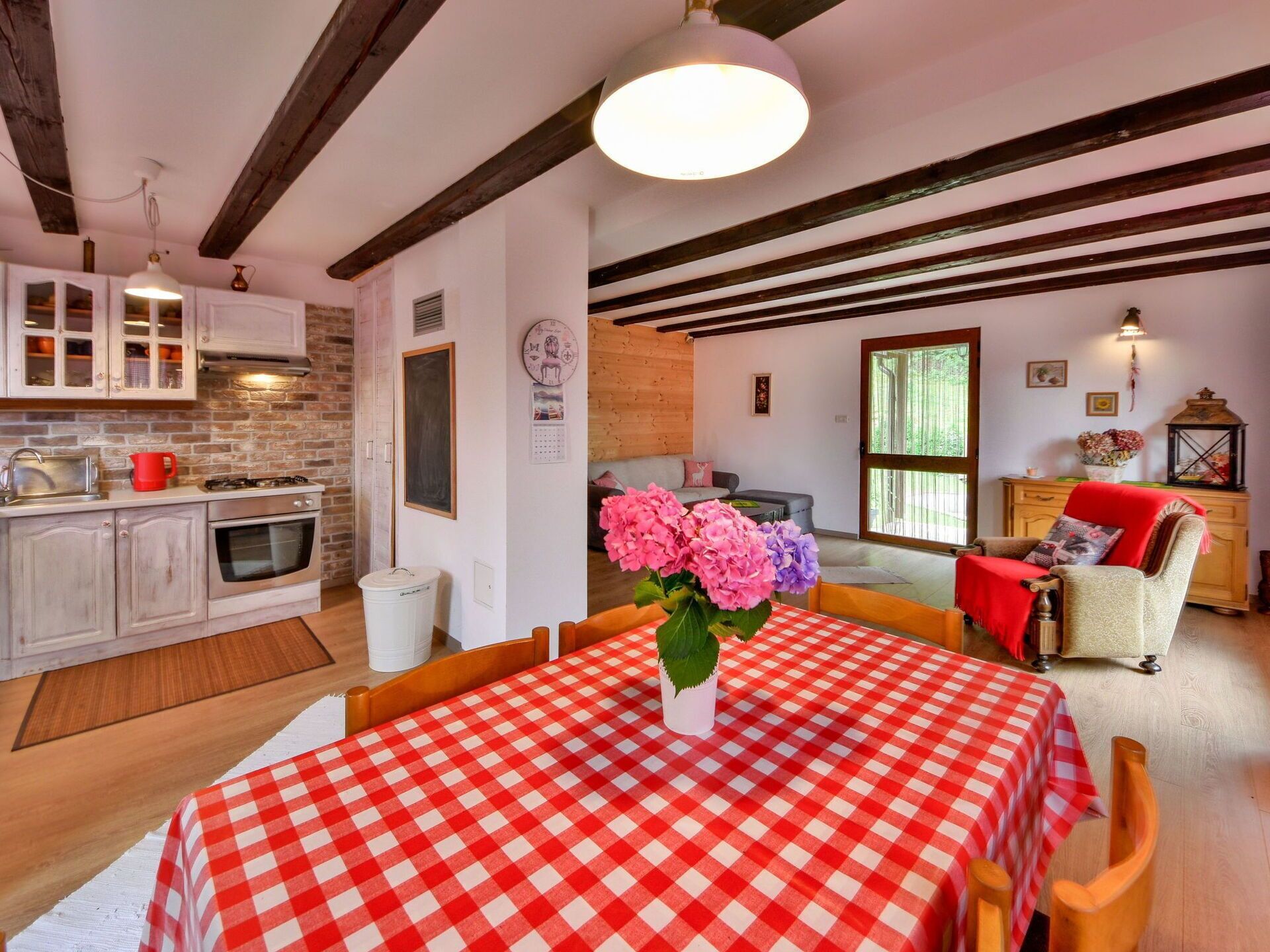 A cozy kitchen with a dining area, featuring a checkered tablecloth, wooden beams, and a large window.