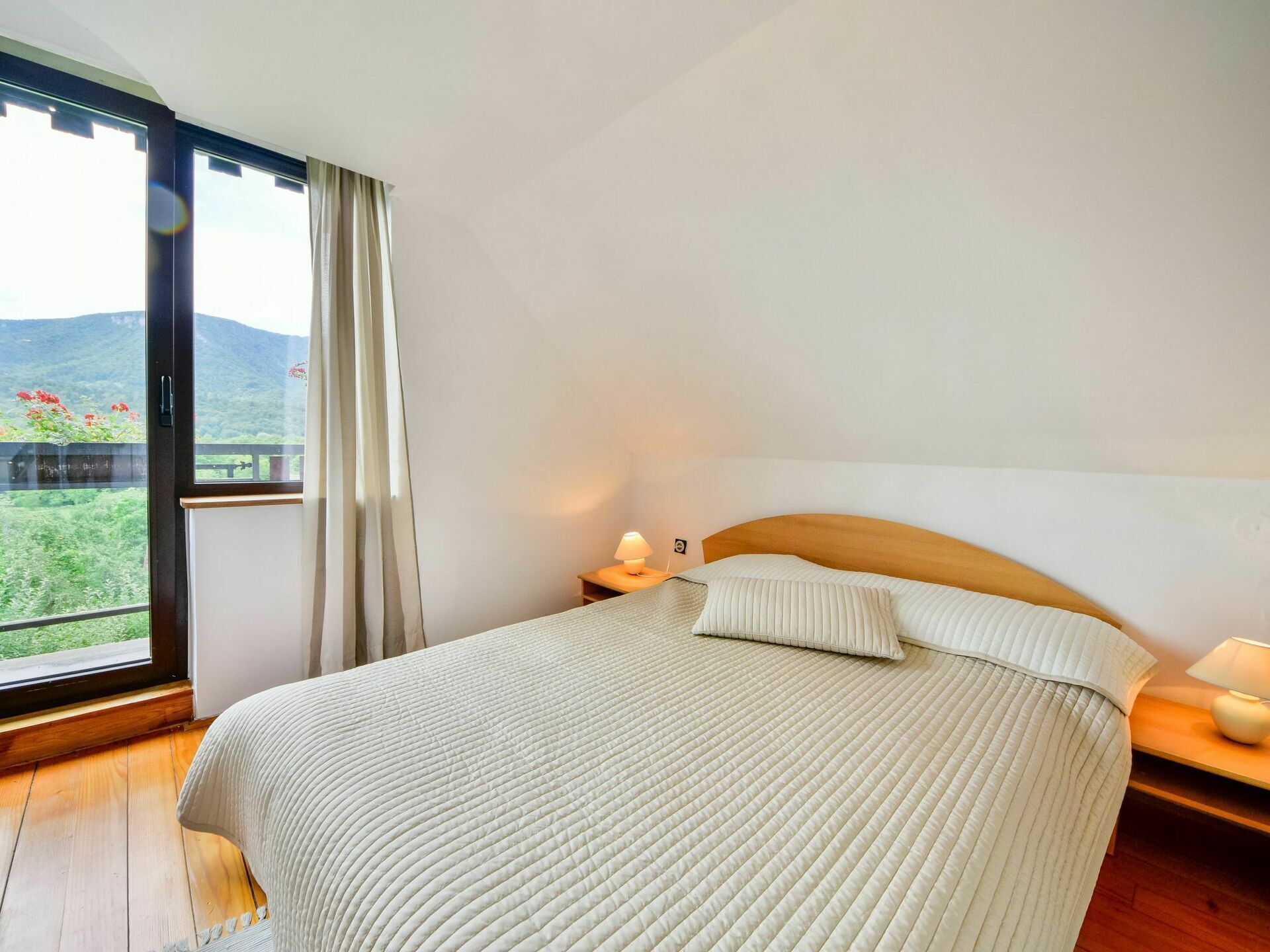 Un dormitorio con una cama grande, una mesita de noche, una lámpara y vistas a un paisaje montañoso a través de un amplio ventanal.