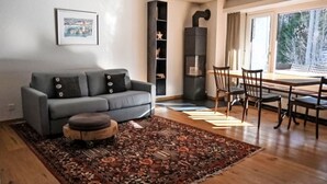 Een moderne woonkamer met een grijze bank, een houten poef, een kamerkleed met een patroon, een eettafel met stoelen en een boekenkast.