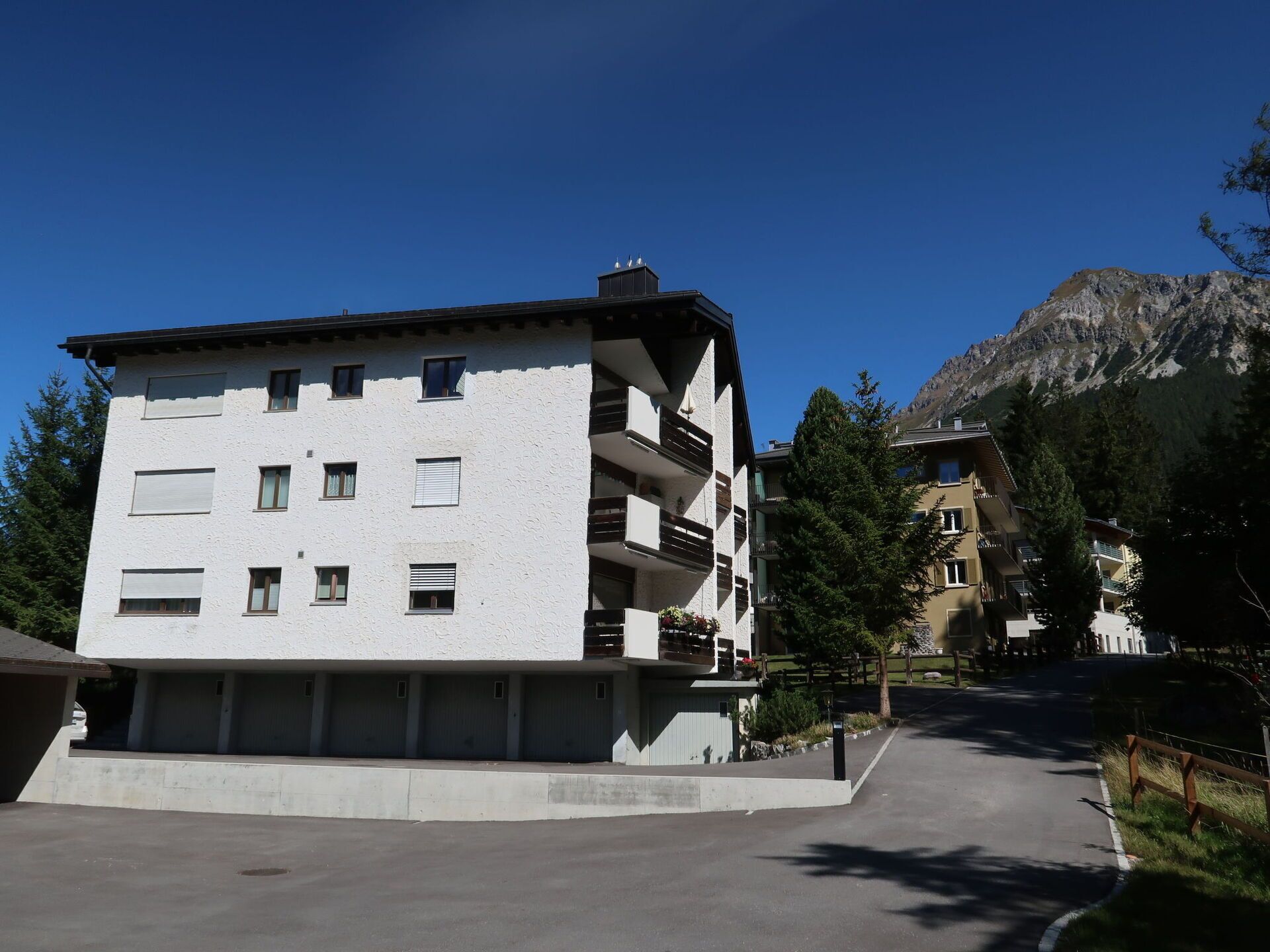 A multi-story building with balconies, a parking area, and a mountain backdrop.