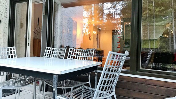 Een terras met een witte tafel en stoelen, een glazen scheiding en uitzicht op het interieur door het glas.