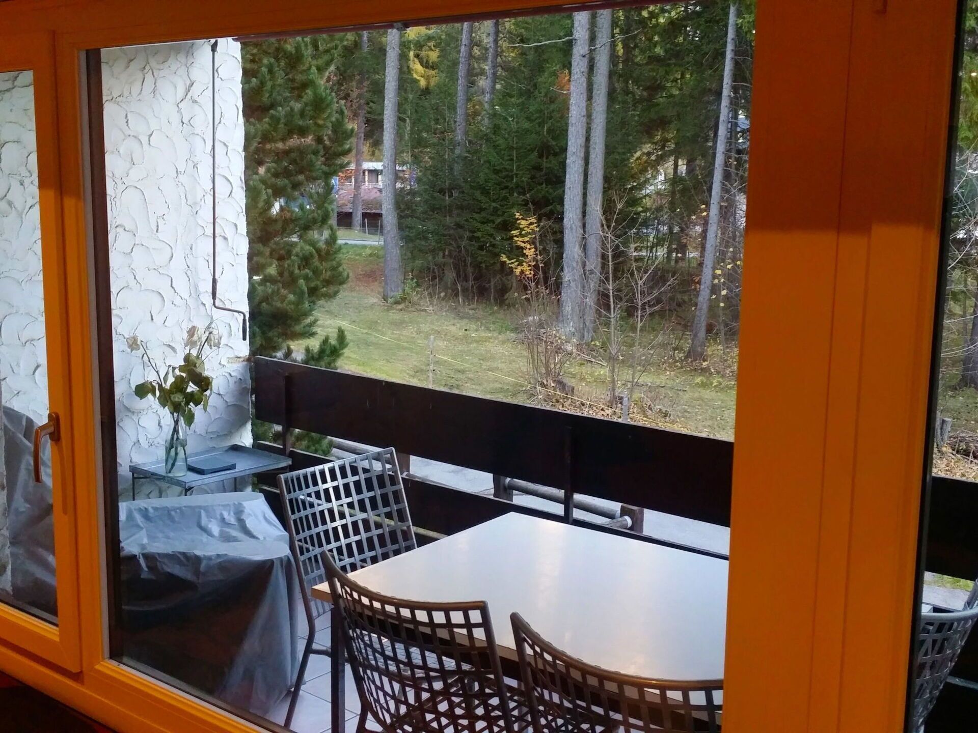 A room with a large window overlooking a forested area, a table with chairs, and a vase with flowers on a side table.
