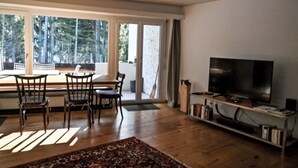 Een woonkamer met een groot raam, een eethoek, een televisie op een dressoir en een boekenkast.