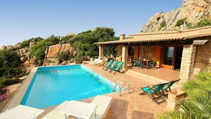 Ein Swimmingpool mit Liegestühlen, eine Terrasse mit Tisch und Stühlen sowie ein Steinhaus mit einem Ziegeldach.